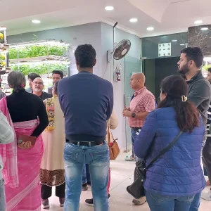 Group of people listening to a presenter in a bright hydroponic plant showroom with vertical racks of leafy greens.",