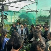 Group of people gathered in a greenhouse, listening to a speaker among rows of plants and hanging pots.